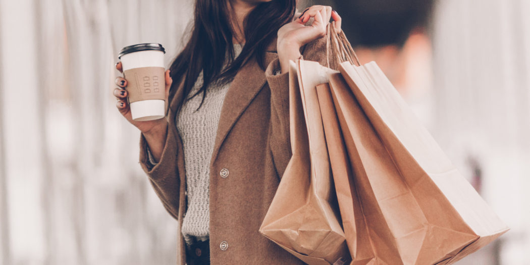 kobieta w beżowym płaszczu z kawą w ręku i z torbami zakupów