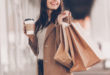 kobieta w beżowym płaszczu z kawą w ręku i z torbami zakupów